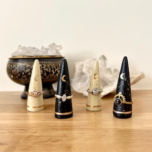 May include: Four decorative ring holders in a row. Two are cream-colored with gold accents and two are black with gold accents. Each holder has a crescent moon design. Rings are displayed on the holders.