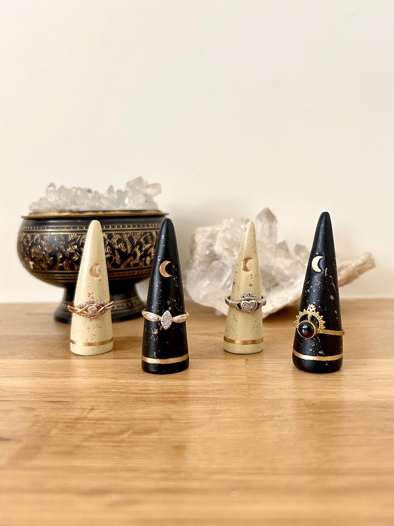 May include: Four decorative ring holders in a row. Two are cream-colored with gold accents and two are black with gold accents. Each holder has a crescent moon design. Rings are displayed on the holders.