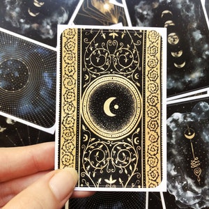 May include: A black and gold tarot card with a crescent moon design. The card has a decorative border with a floral pattern. The card is surrounded by other cards with celestial designs.