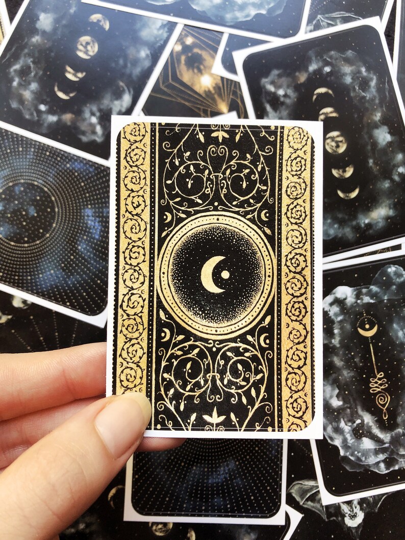 May include: A black and gold tarot card with a crescent moon design. The card has a decorative border with a floral pattern. The card is surrounded by other cards with celestial designs.