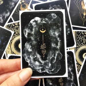 May include: A black and gold card with a white cloud design and a gold crescent moon and geometric symbol. The card is part of a set of cards with a celestial theme.