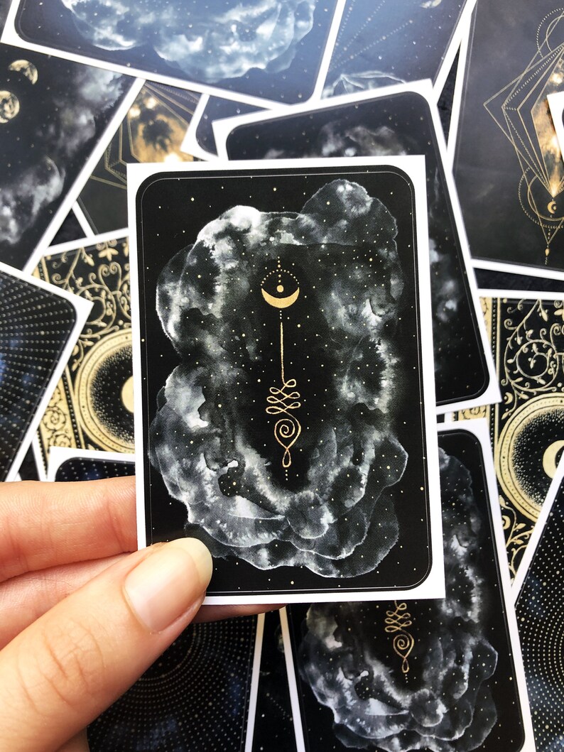 May include: A black and gold card with a white cloud design and a gold crescent moon and geometric symbol. The card is part of a set of cards with a celestial theme.