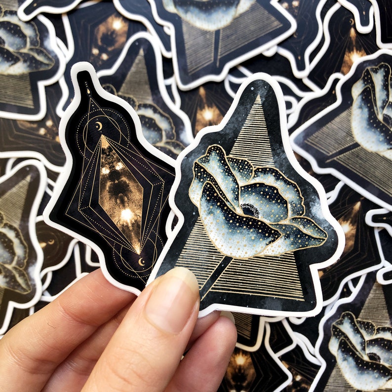 K&ouml;nnte beinhalten: Zwei schwarze und goldene geometrische Aufkleber mit wei&szlig;em Rand. Ein Aufkleber zeigt ein schwarzes und goldenes geometrisches Design mit einer wei&szlig;en Mohnblume in der Mitte. Der andere Aufkleber zeigt ein schwarzes und goldenes geometrisches Design mit einer wei&szlig;en Mohnblume in der Mitte.