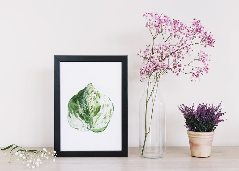 May include: A framed watercolor painting of a green and white leaf, displayed on a table with a vase of pink flowers and a potted plant with purple flowers.
