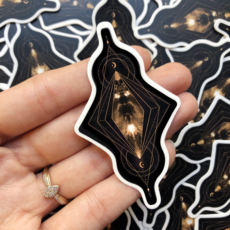 K&ouml;nnte beinhalten: Ein geometrischer Aufkleber in Schwarz und Gold mit einem himmlischen Design. Der Aufkleber zeigt eine Diamantform mit einem Sternenhimmel im Inneren, umgeben von einem Kreis mit einer Mondsichel und Sternen. Der Aufkleber ist perfekt, um Ihrem Laptop, Ihrer Wasserflasche oder Ihrem Planer einen Hauch von himmlischer Magie zu verleihen.