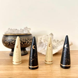 May include: Four decorative cone-shaped objects with a celestial theme. Two are black with gold accents and a crescent moon design, and two are cream-colored with gold accents and a crescent moon design. The objects are displayed on a wooden surface.