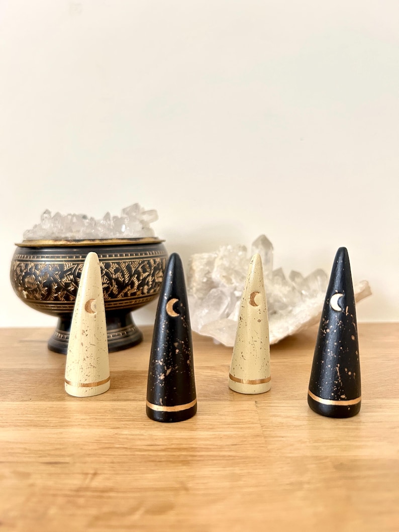 May include: Four decorative cone-shaped objects with a celestial theme. Two are black with gold accents and a crescent moon design, and two are cream-colored with gold accents and a crescent moon design. The objects are displayed on a wooden surface.