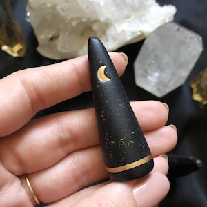 May include: Black, cone-shaped decorative object with a gold crescent moon and a gold band near the base. The object has a slightly textured surface and is held in a hand. Crystals are in the background.