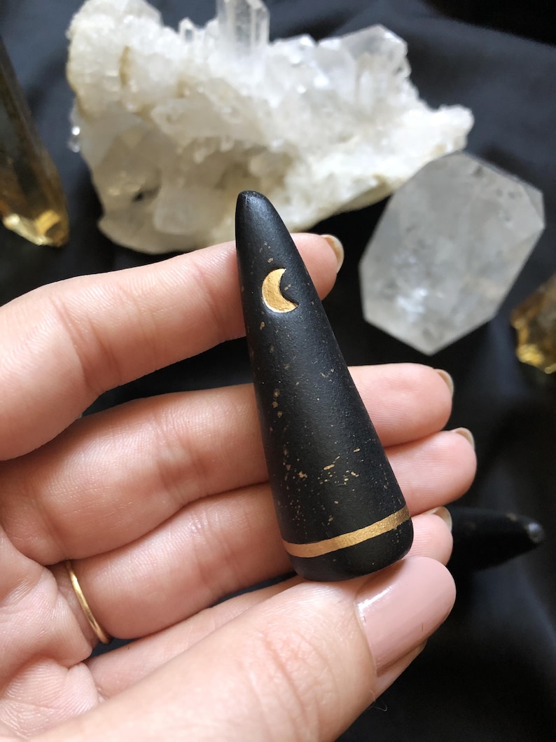 May include: Black, cone-shaped decorative object with a gold crescent moon and a gold band near the base. The object has a slightly textured surface and is held in a hand. Crystals are in the background.