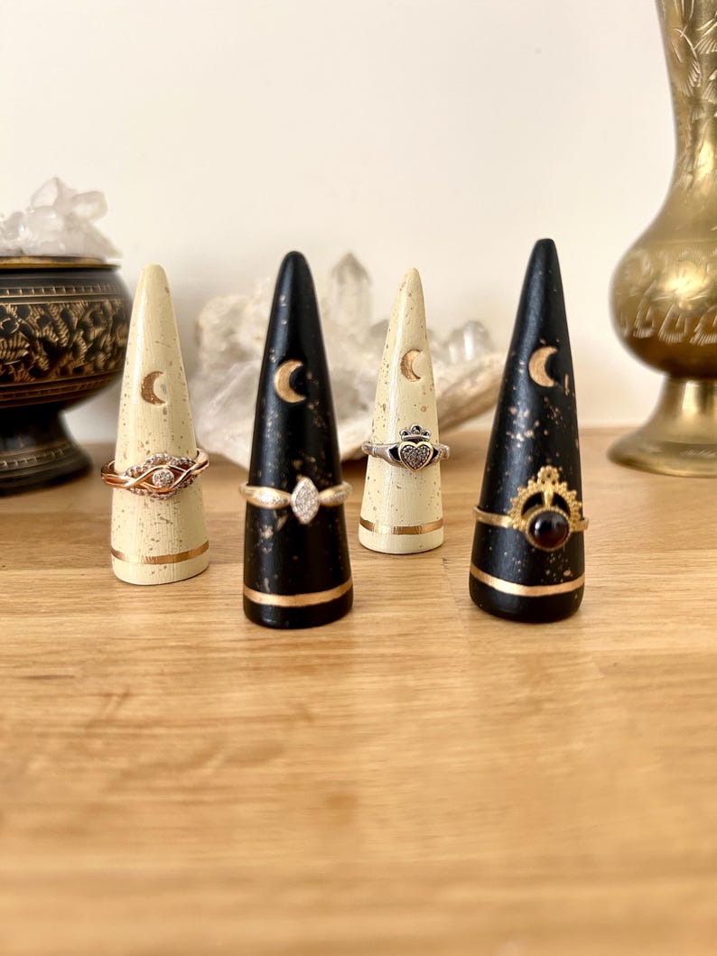 May include: Four decorative ring holders in a row. Two are black with gold accents and two are cream-colored. Each holder has a crescent moon design and a gold band. Rings of various designs are displayed on the holders.