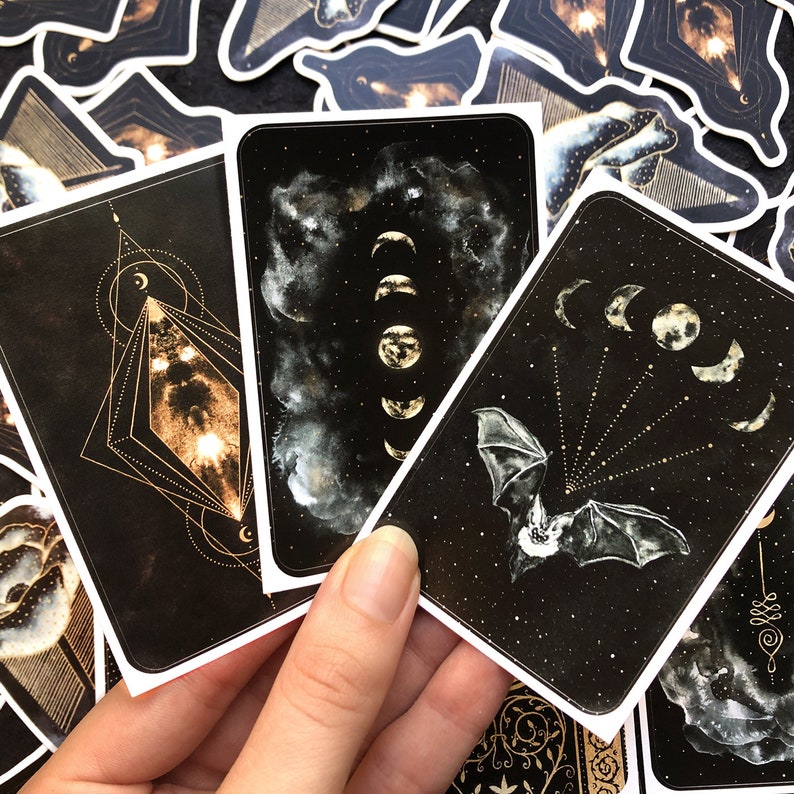 May include: Three black tarot cards with gold accents. The cards feature a geometric design, a crescent moon and stars, and a bat with a crescent moon above it.