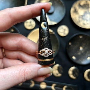 May include: A gold and diamond ring is displayed on a black ring holder with a gold band and moon cutout. The ring holder is surrounded by other celestial-themed decor with gold accents.