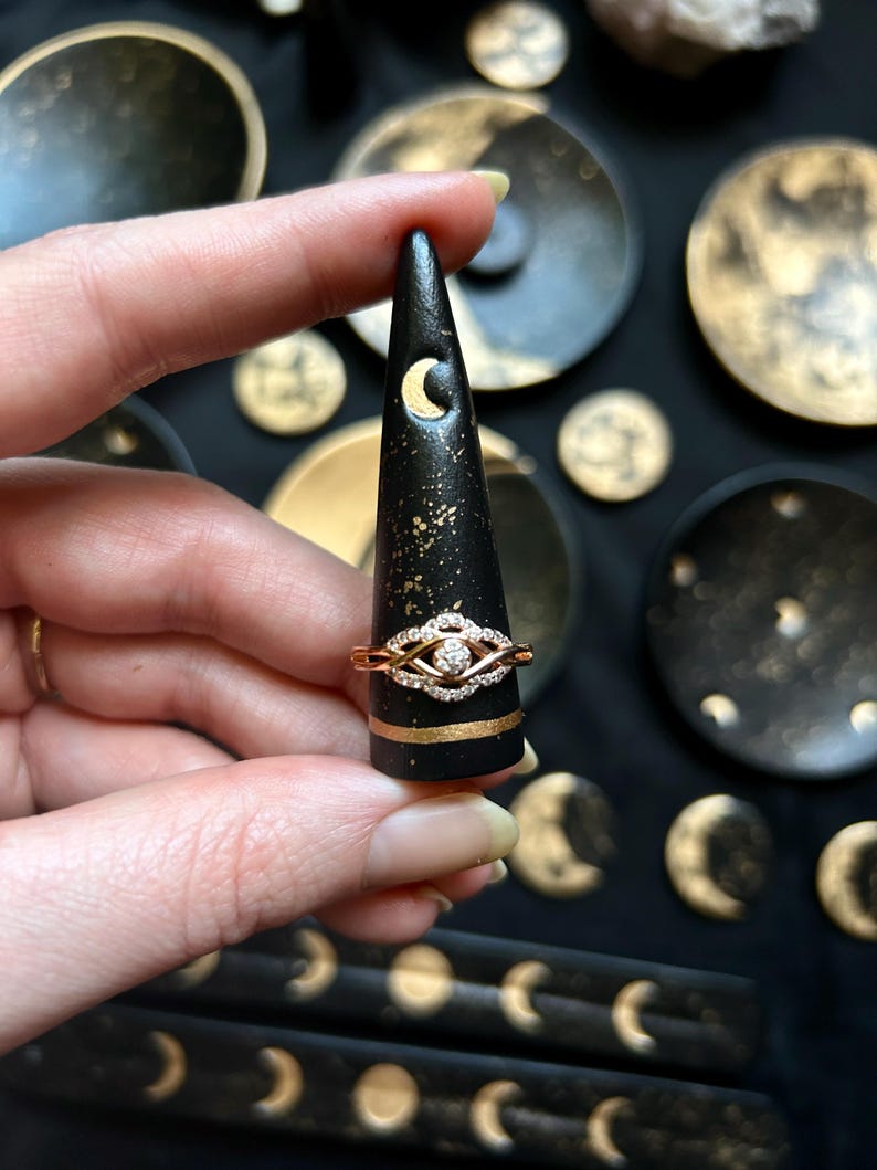 May include: A gold and diamond ring is displayed on a black ring holder with a gold band and moon cutout. The ring holder is surrounded by other celestial-themed decor with gold accents.