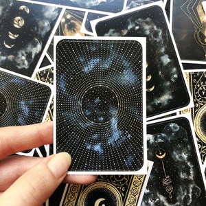 May include: A black and gold tarot card with a celestial design. The card features a circular pattern of gold dots and a blue nebula in the center. The card is surrounded by a gold border with a crescent moon design.