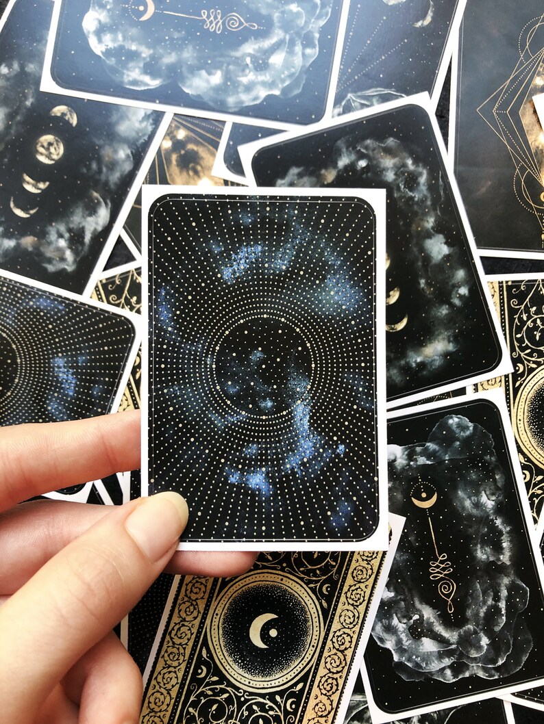 May include: A black and gold tarot card with a celestial design. The card features a circular pattern of gold dots and a blue nebula in the center. The card is surrounded by a gold border with a crescent moon design.