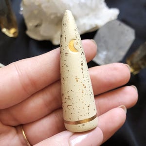 May include: A cream-colored, cone-shaped incense holder with gold accents. The holder features a gold crescent moon design and gold speckles. A gold band encircles the base. The holder is held in a hand.