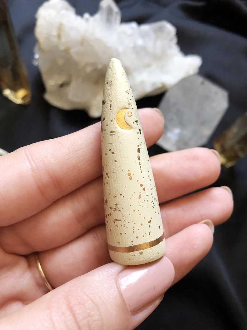 May include: A cream-colored, cone-shaped incense holder with gold accents. The holder features a gold crescent moon design and gold speckles. A gold band encircles the base. The holder is held in a hand.