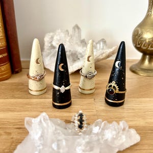 May include: A collection of ring holders in cream and black with gold accents. Each holder is cone-shaped and features a crescent moon design. Rings with various designs are displayed on the holders, with a ring also resting on a clear crystal.