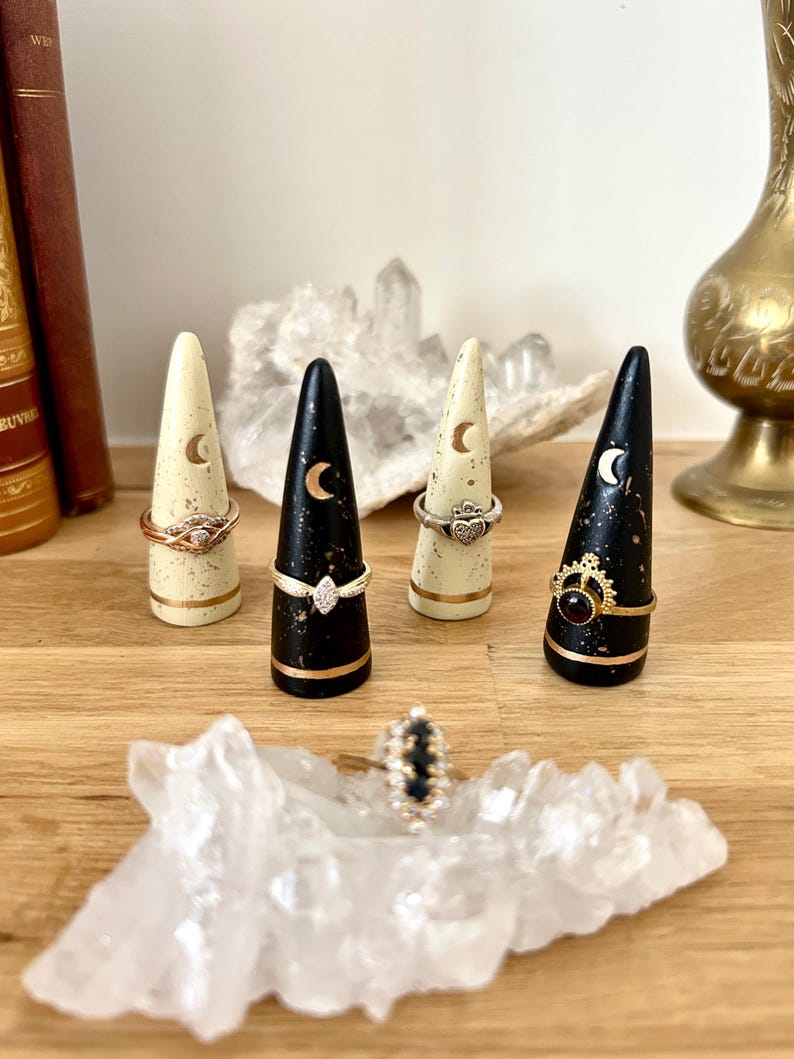 May include: A collection of ring holders in cream and black with gold accents. Each holder is cone-shaped and features a crescent moon design. Rings with various designs are displayed on the holders, with a ring also resting on a clear crystal.