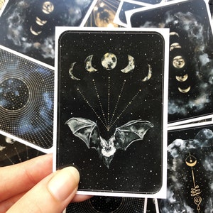 May include: A black card with a white bat flying in front of a crescent moon and stars. The bat is facing forward with its wings spread out. The moon is in the center of the card and is surrounded by five crescent moons.
