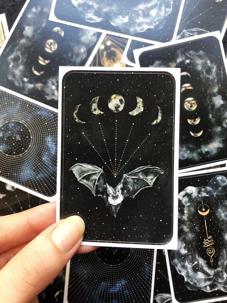 May include: A black card with a white bat flying in front of a crescent moon and stars. The bat is facing forward with its wings spread out. The moon is in the center of the card and is surrounded by five crescent moons.