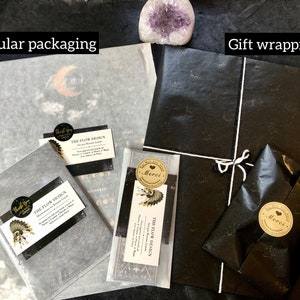 May include: Two images showing packaging options for a print. The first image shows a print wrapped in white tissue paper with a black and gold sticker that says "The Flow Design". The second image shows a print wrapped in black tissue paper with a gold sticker that says "Merci".