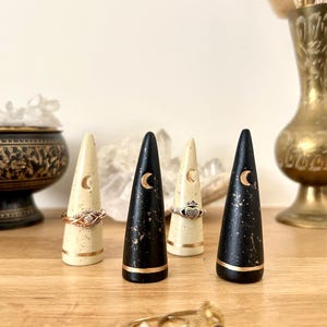 May include: A collection of ring holders in cream and black with gold accents. Each holder is cone-shaped and features a crescent moon design. Rings are displayed on the holders, with a gold and diamond ring in the foreground.