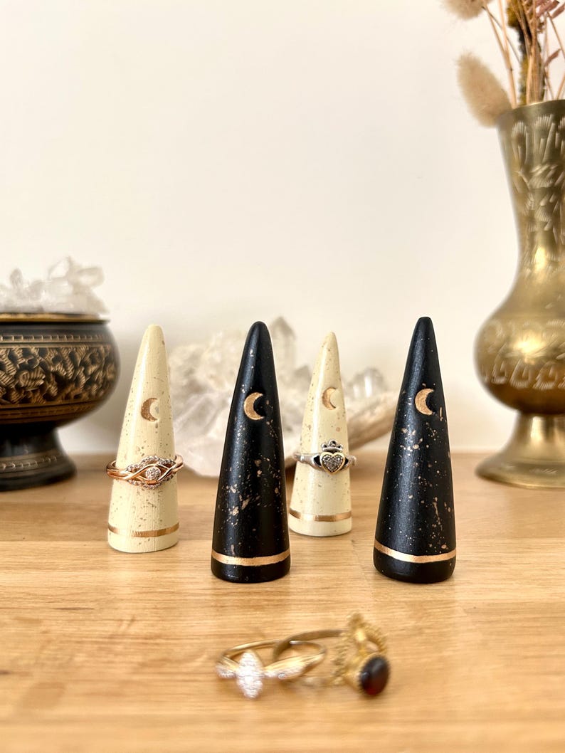 May include: A collection of ring holders in cream and black with gold accents. Each holder is cone-shaped and features a crescent moon design. Rings are displayed on the holders, with a gold and diamond ring in the foreground.