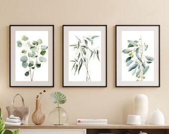 Può includere: Tre stampe botaniche incorniciate con illustrazioni ad acquerello di eucalipto e rami d'ulivo. Le stampe hanno cornici nere e sono esposte sopra una consolle in legno con oggetti decorativi e una pianta in vaso. La tavolozza dei colori è neutra con accenti verdi.
