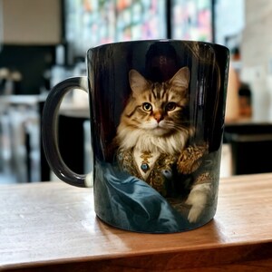 Puede incluir: Una taza de cerámica negra con asa presenta un retrato de un gato de pelo largo que lleva una túnica azul y dorada. El gato tiene ojos verdes y una expresión seria.