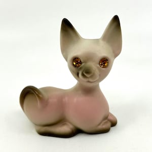 May include: A small ceramic cat figurine with a light pink and cream body, dark brown shading on the ears, tail, and paws, and two faceted amber-colored rhinestone eyes. The cat is in a seated position with its tail curled upward.
