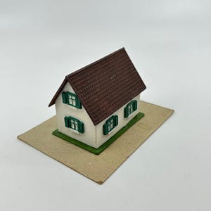 Puede incluir: Una casa modelo en miniatura con paredes de color blanco roto, un tejado a dos aguas de color marrón oscuro texturizado y contraventanas verdes. La estructura se asienta sobre una base rectangular e incluye varias ventanas con marcos blancos.