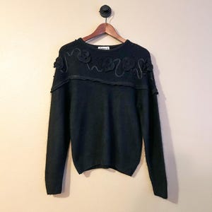 Op de afbeelding: Zwarte trui met bloemversieringen en kraalaccenten. De trui met ronde hals heeft een decoratief ontwerp over de schouders met driedimensionale bloemen en kleine kralen. De trui met lange mouwen hangt aan een houten hanger.