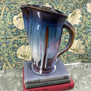 May include: A ceramic pitcher featuring a glossy glaze with blended shades of deep blue, purple, and white, with brown accents along the rim and handle. The pitcher has a wide spout and a curved handle, resting on a stack of two hardcover books.