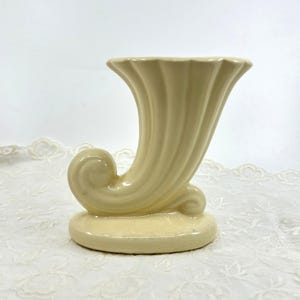 Puede incluir: Un jarrón de cerámica color crema con forma de cuerno de la abundancia, acabado brillante y estrías verticales. El recipiente tiene una abertura ancha y acampanada en la parte superior y una base curva en espiral sobre un pedestal.