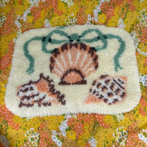 May include: A rectangular, cream-colored rug with a seashell design. The rug features a large, detailed seashell in the center, flanked by two smaller shells. A green bow is at the top. The shells are in shades of peach and brown.