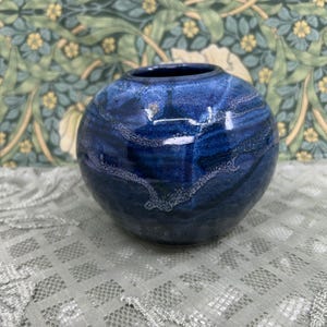 May include: A small, spherical ceramic vase featuring a deep blue glaze with swirling, lighter blue and white textured patterns across its surface. The vase has a smooth, rounded body and a circular opening at the top, showcasing a glossy finish.