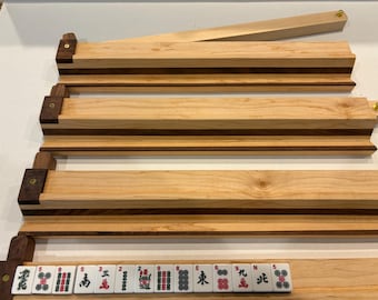 May include: Four wooden Mahjong tile racks featuring a two-tone design with light maple and dark walnut wood. Each rack has a grooved ledge for holding tiles, a dark wood end block with a gold-colored screw, and one rack displays a row of tiles.