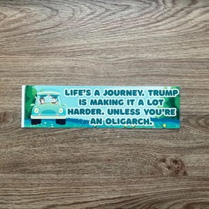 Puede incluir: Una pegatina rectangular con el texto "LIFE'S A JOURNEY. TRUMP IS MAKING IT A LOT HARDER. UNLESS YOU'RE AN OLIGARCH." Un coche turquesa con dos personas está en el lado izquierdo de la pegatina.