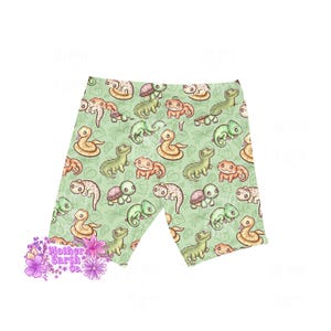 Op de afbeelding: Lichtgroene biker shorts met een herhaald patroon van cartoon slangen, schildpadden en hagedissen. De shorts hebben een verscheidenheid aan kleuren, waaronder bruin, groen en oranje, tegen een lichtgroene achtergrond met blad details. De tekst "Mother Earth Co." is zichtbaar.