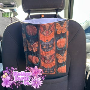 May include: A black car trash bag with a Halloween-themed pattern. The pattern includes orange pumpkins, bats, butterflies, and black cats. The bag is hanging from the headrest of a car seat.