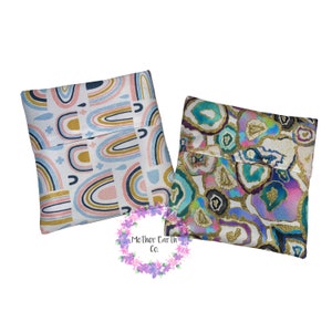 Puede incluir: Dos bolsas de tela para aperitivos, una con un patrón de arcoíris y la otra con un patrón de ágata azul y púrpura. Ambas bolsas tienen un fondo blanco y detalles dorados.