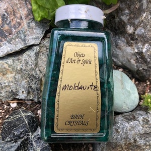 May include: A clear glass jar filled with green bath crystals. The jar has a gold label that reads "Objects d'Art & Spirit Moldavite Bath Crystals".