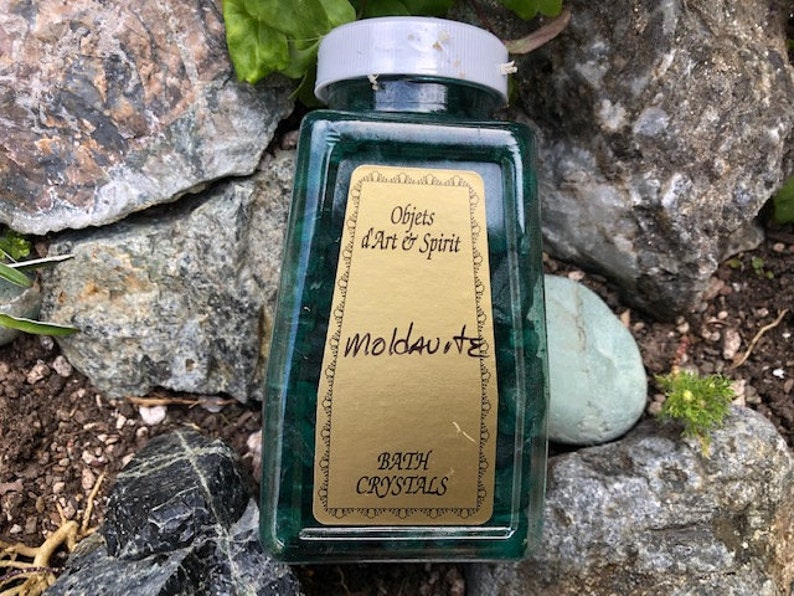 May include: A clear glass jar filled with green bath crystals. The jar has a gold label that reads "Objects d'Art & Spirit Moldavite Bath Crystals".