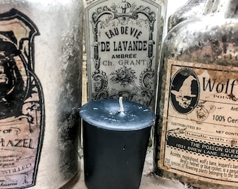 May include: Three glass bottles with vintage labels, one with a black and white illustration of a witch, one with a black and white illustration of a wolf, and one with a black and white illustration of a flower. A black candle sits in front of the bottles.
