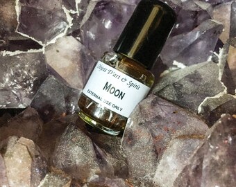 May include: A small glass bottle with a black cap, filled with a brown liquid. The label reads "Objets D'art & Spirit" and "MOON" in white text. The label also states "EXTERNAL USE ONLY". The bottle is sitting on a bed of purple amethyst crystals.