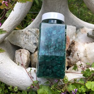 May include: A glass jar filled with teal green and black crushed crystals. The jar has a white lid and is sitting on a white rock.