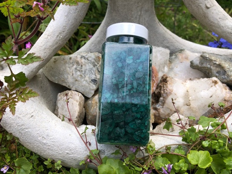 May include: A glass jar filled with teal green and black crushed crystals. The jar has a white lid and is sitting on a white rock.