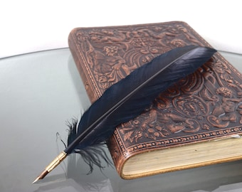 May include: A dark black feather quill pen with a metallic gold-toned nib resting diagonally across a rectangular brown leather-bound journal featuring an embossed relief pattern of figures and intricate scrollwork on the front cover.