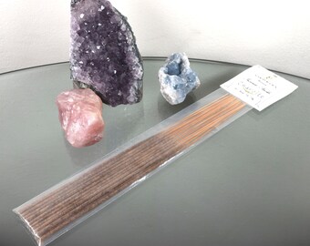 Può includere: Un pacchetto di bastoncini di incenso marroni con un'etichetta bianca che recita "Sandalwood" e "Incense Sticks". Il pacchetto è su una superficie di vetro con tre cristalli: un grande ametista, un piccolo quarzo rosa e un cristallo blu.